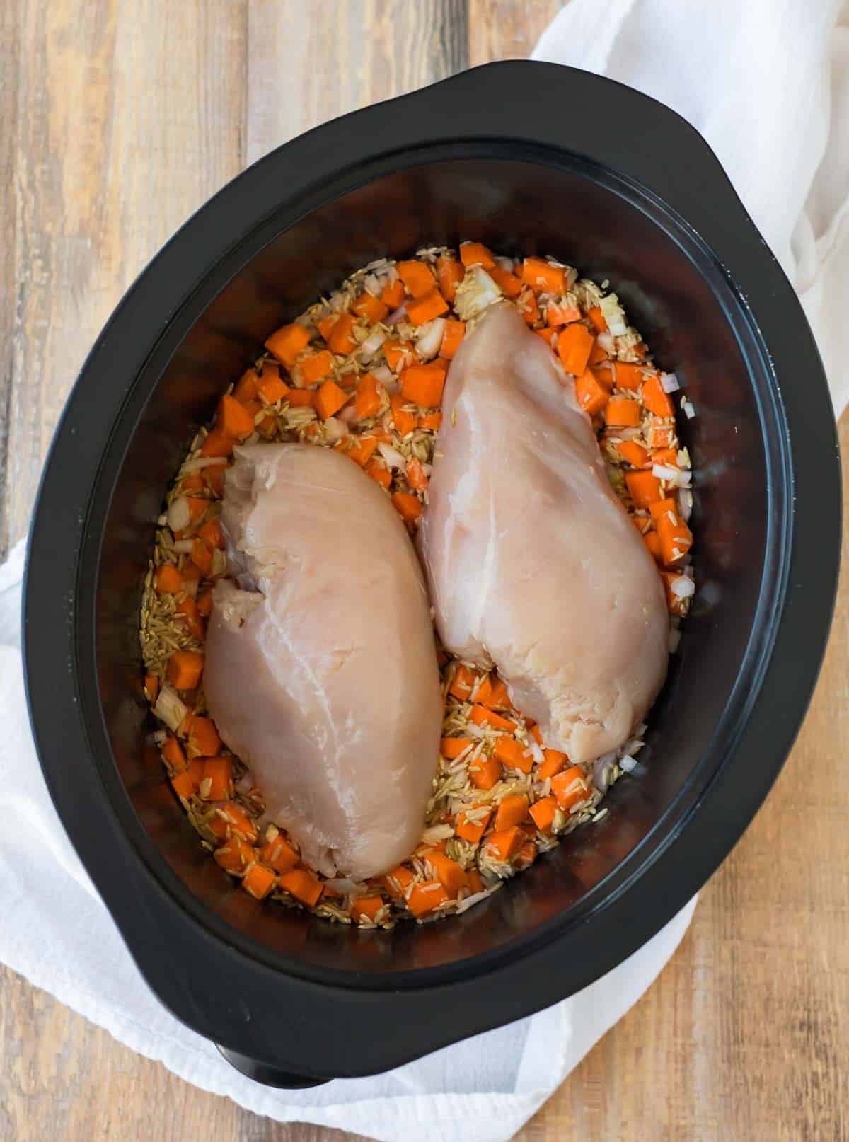 Two pieces of raw poultry on a bed of vegetables and grains in a slow cooker