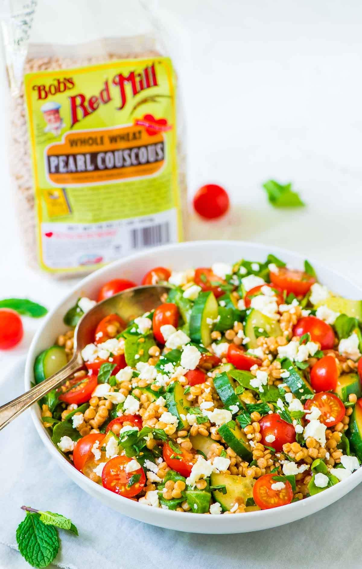 Tomato, Cucumber, and Feta Pearl Couscous Salad in a white bowl, with a bag of Bob's Red Mill pearl couscous behind it
