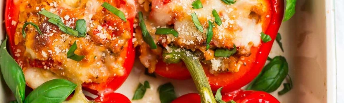 A pan of healthy Italian stuffed peppers; red bell peppers filled with ground chicken, tomatoes, and whole grains, then topped with basil and cheese.