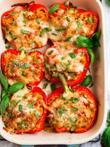 A pan of healthy Italian stuffed peppers; red bell peppers filled with ground chicken, tomatoes, and whole grains, then topped with basil and cheese.