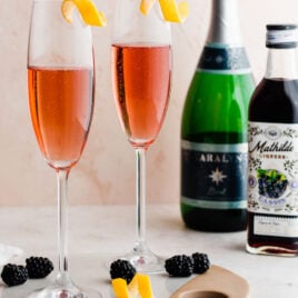 Two Kir Royale drinks garnished with a lemon twist and blackberries and the ingredients, champagne and cassis liqueur in the background